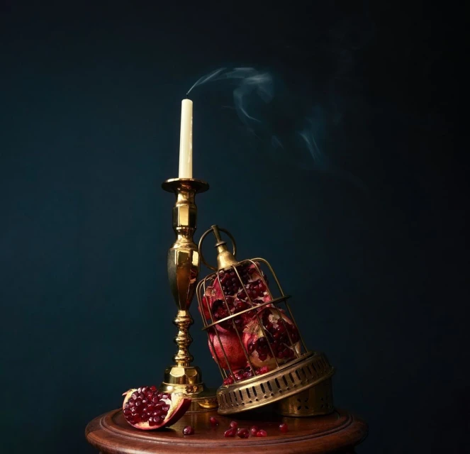 Still life photograph of a pomegranate in a cage, extinguished candle on a brass candlestick, and pomegranate seeds on a wooden table.