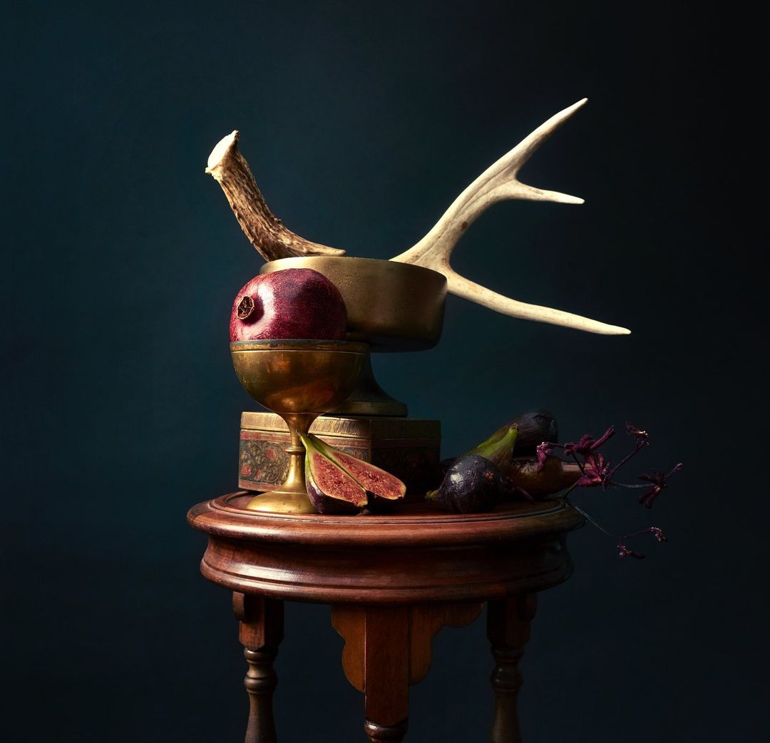 Still life photography featuring a pomegranate, figs, deer antlers, and antique brass objects on a wooden pedestal against a dark backdrop.