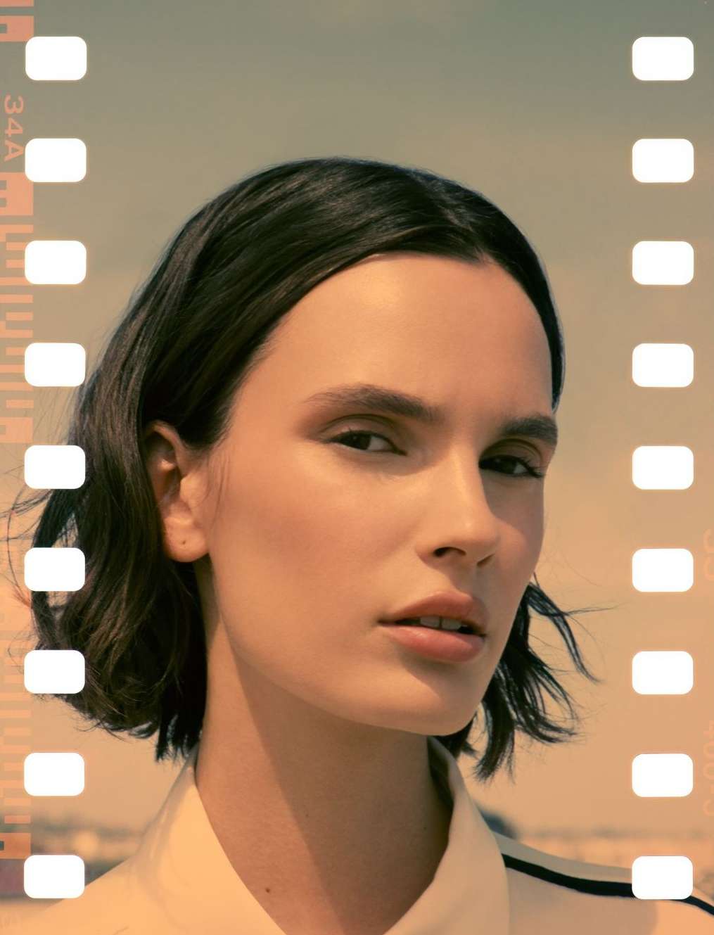 Woman with bob haircut poses indoors with film border. Close-up headshot with neutral expression, warm lighting, and minimalist aesthetic.