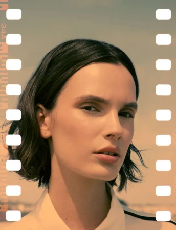 Woman with bob haircut poses indoors with film border. Close-up headshot with neutral expression, warm lighting, and minimalist aesthetic.