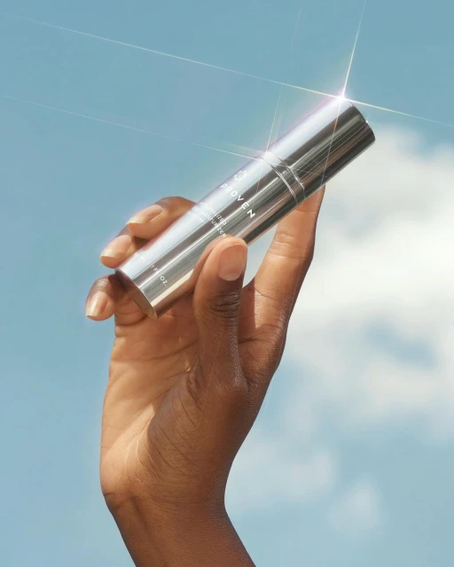 A dark-skinned hand holds a silver-colored skincare product against a bright blue sky with a few faint clouds. The packaging is minimalist.