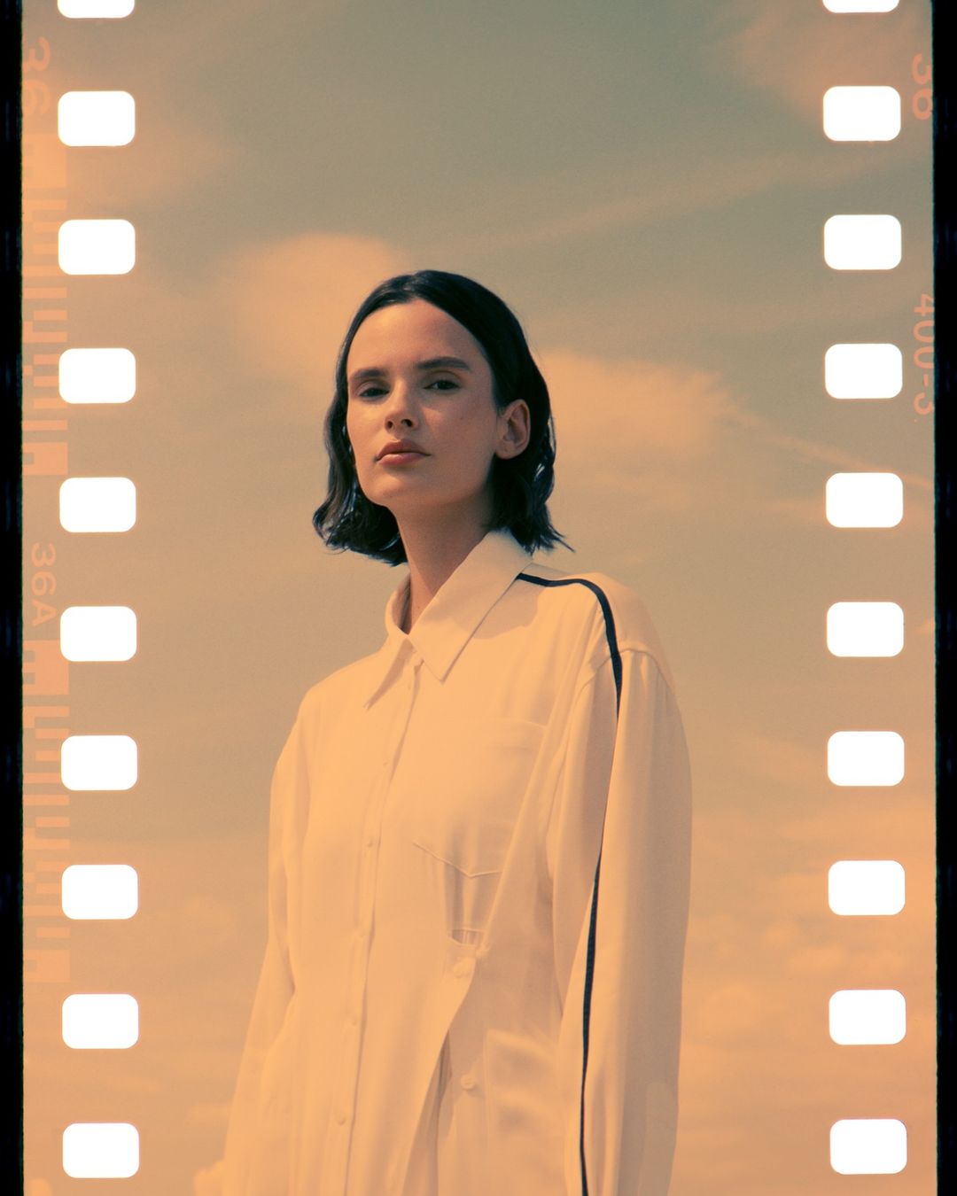 Woman with short dark hair posing outdoors in a white shirt with blue trim against a hazy sky framed by film strip border.