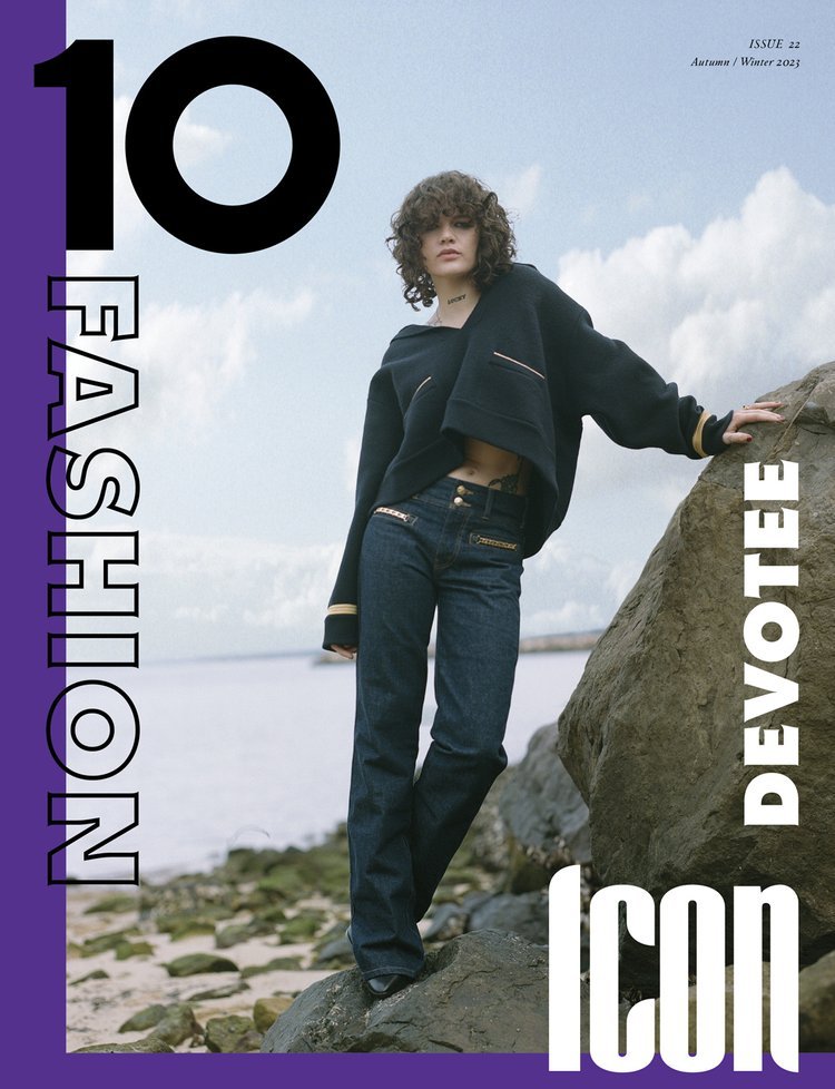 Magazine cover shot of curly-haired model wearing a black jacket and jeans, standing near rocks with a cloudy sky and water backdrop.