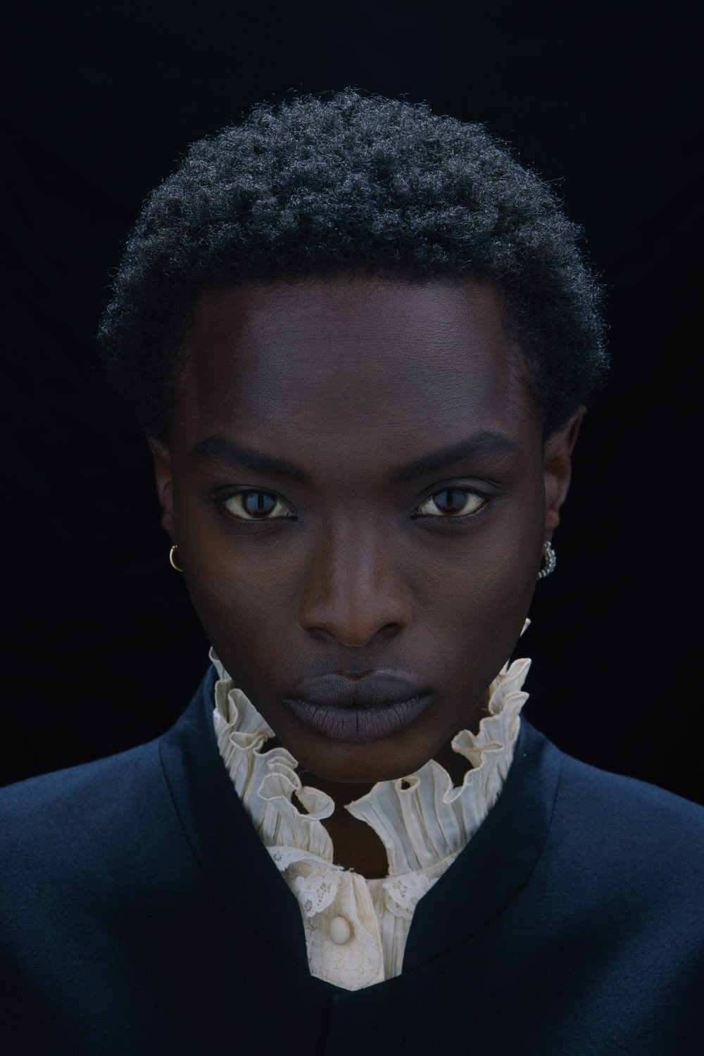 Close-up portrait of an African American woman with short curly hair, wearing a dark blue jacket and white frilled collar, against black background.