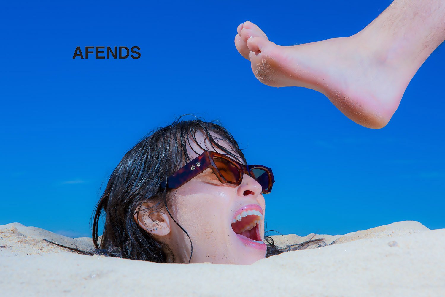 Person buried in sand with sunglasses, blue sky background, and foot. Summer beach scene with a playful, surreal composition.