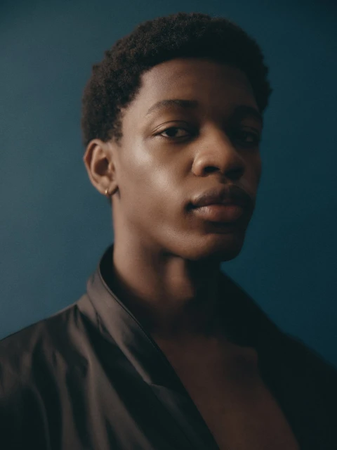 Close-up portrait of a young Black man in a dark robe against a dark blue background. He has short natural hair.