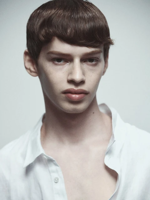 Portrait of a young adult man with dark hair and a white shirt, looking at the camera against a neutral light background.