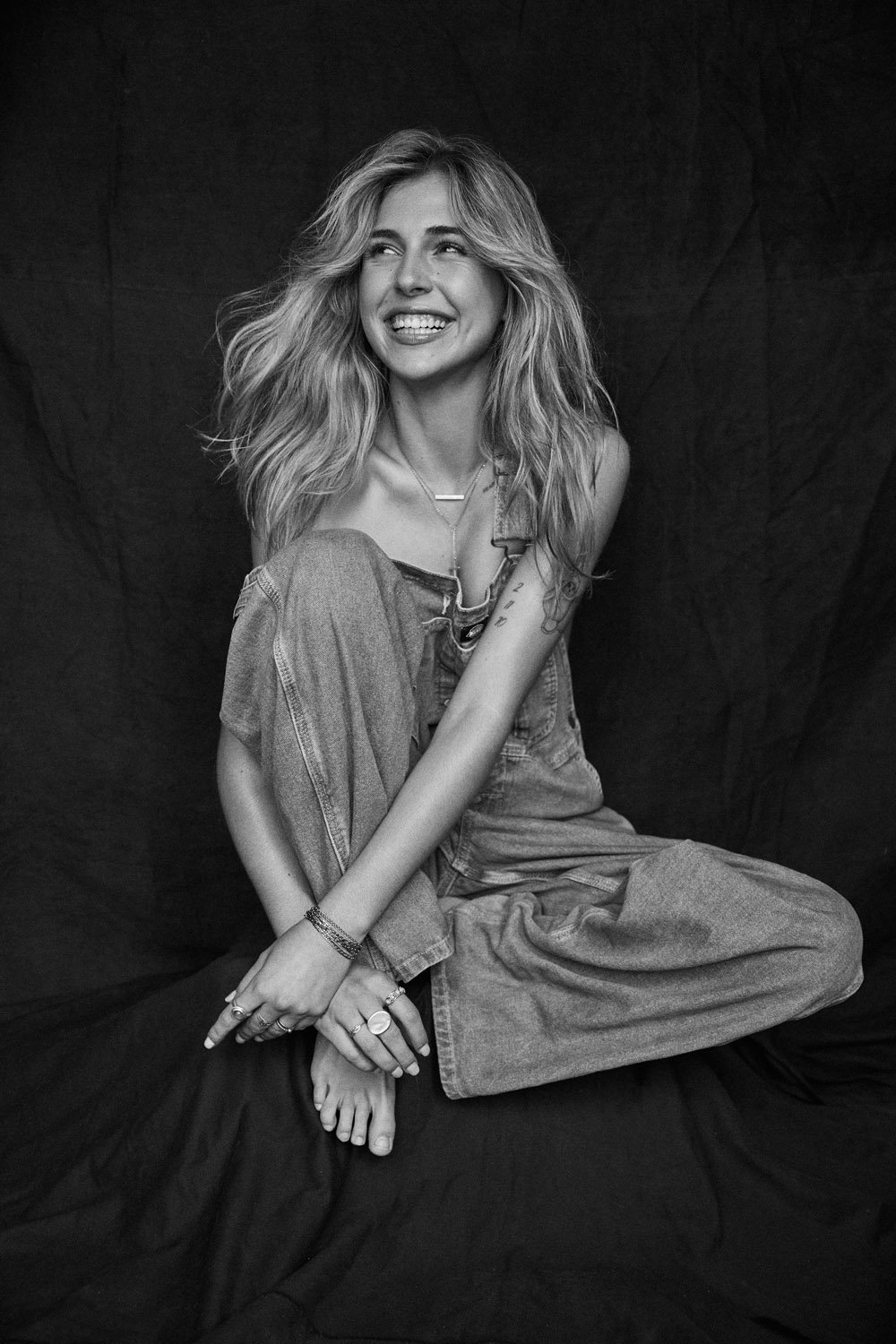 Portrait of a smiling woman with long blonde hair sitting in denim overalls against a dark backdrop, hands folded in her lap.