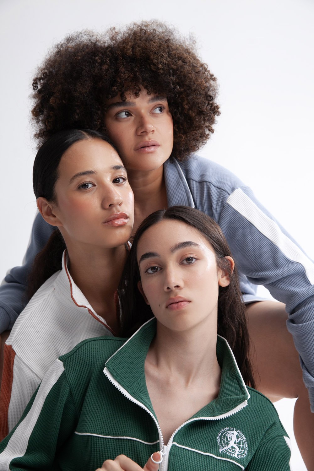 Three female models in tracksuits pose in a fashion shot in a bright photo studio. The models wear sporty tops with white trim details.