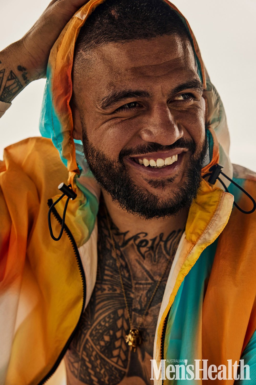 Smiling man with tribal tattoos and a colorful hooded jacket, captured in a close-up studio shot for Men's Health magazine.