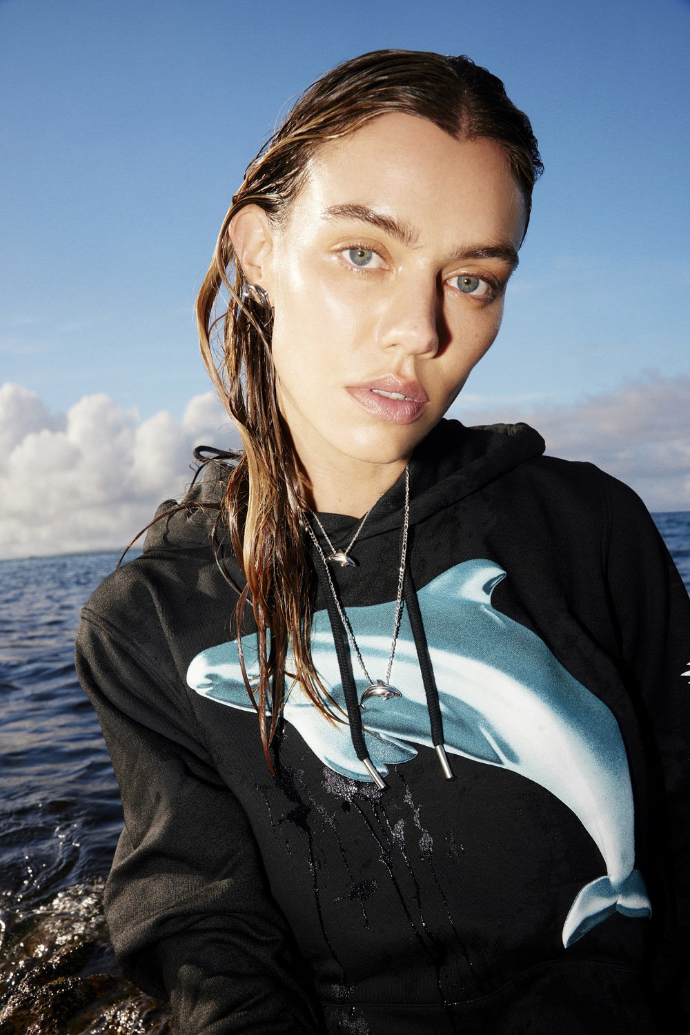 Woman in a black hoodie with a dolphin graphic poses outdoors. Blue sky and ocean water create a fresh, natural backdrop.