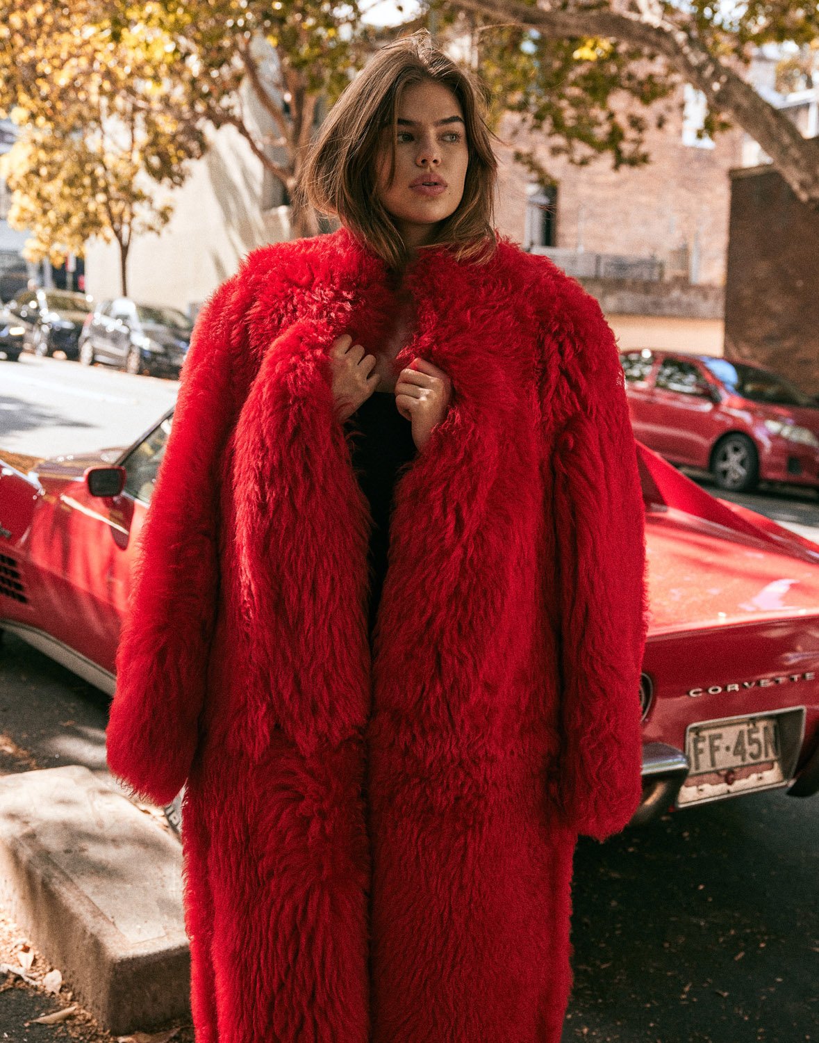 Woman in a red faux fur coat stands beside a vintage red Corvette in an urban setting, capturing a blend of fashion and automotive.
