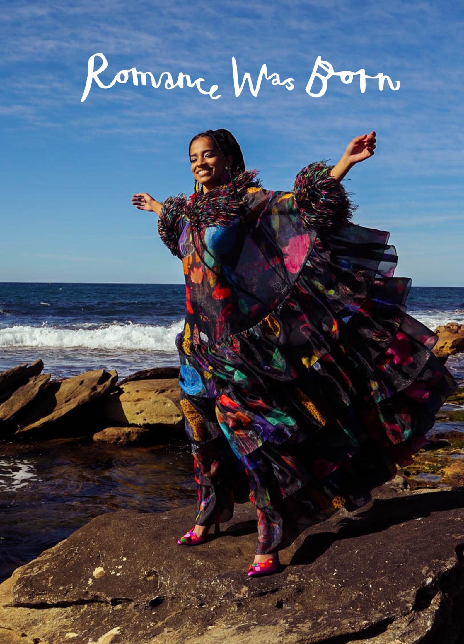 A woman in a flowing dress poses on rocks by the ocean under a clear blue sky, exuding a vibrant and carefree atmosphere.