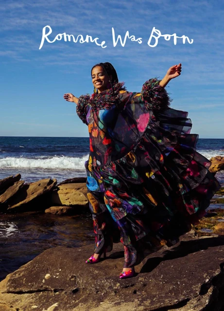 A woman in a flowing dress poses on rocks by the ocean under a clear blue sky, exuding a vibrant and carefree atmosphere.