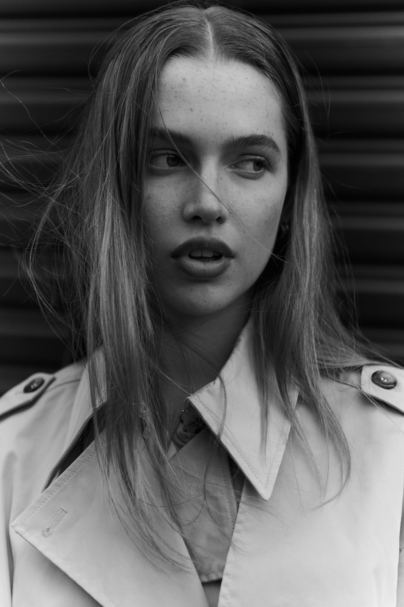 Portrait of a young woman in a beige trench coat against a dark background, with long blonde hair blowing in the wind in black and white.