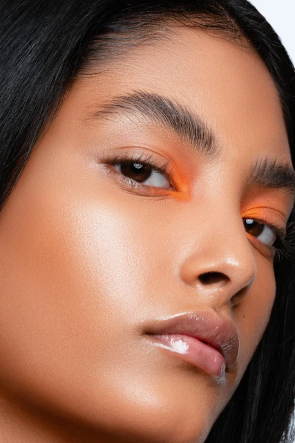 Close-up of a woman's face featuring orange eyeshadow and glossy lips, highlighting her skin tone and dark hair.