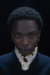 Portrait of a Black model with short textured hair wearing a high-collared ruffled lace shirt and dark jacket against a black background.