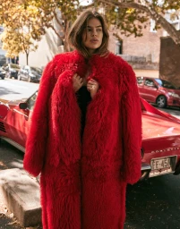 A woman wearing a vibrant red faux fur coat standing in front of a parked red sports car on a city street.