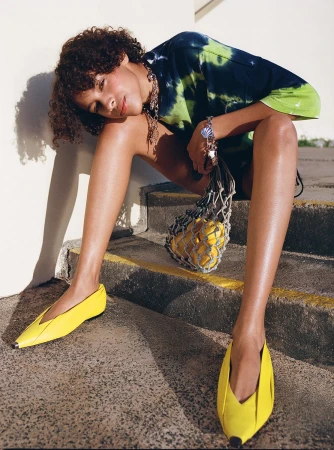 Model with curly hair wearing tie dye t-shirt and yellow shoes sitting on outdoor stone steps holding string bag with lemons