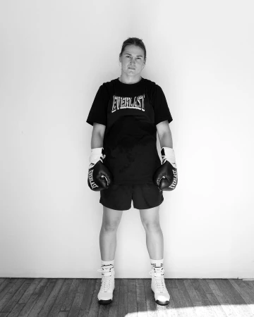 Black and white portrait of a woman in boxing attire, including gloves and boots, standing against a light wall with wood flooring.