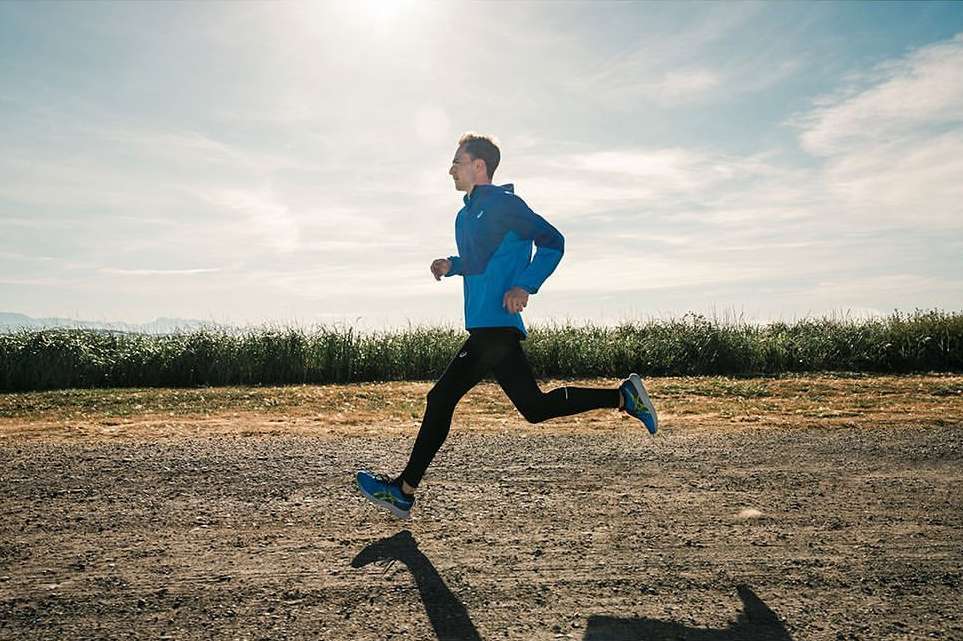 A male runner in a blue jacket and black leggings is captured in mid-stride on a dirt path with grassy fields and a bright sky.