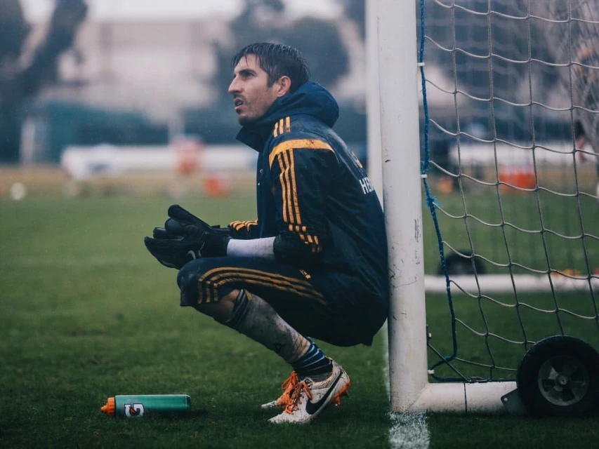 Male soccer goalie wearing protective gloves crouches on a grassy field next to a net with a sports bottle at his feet.