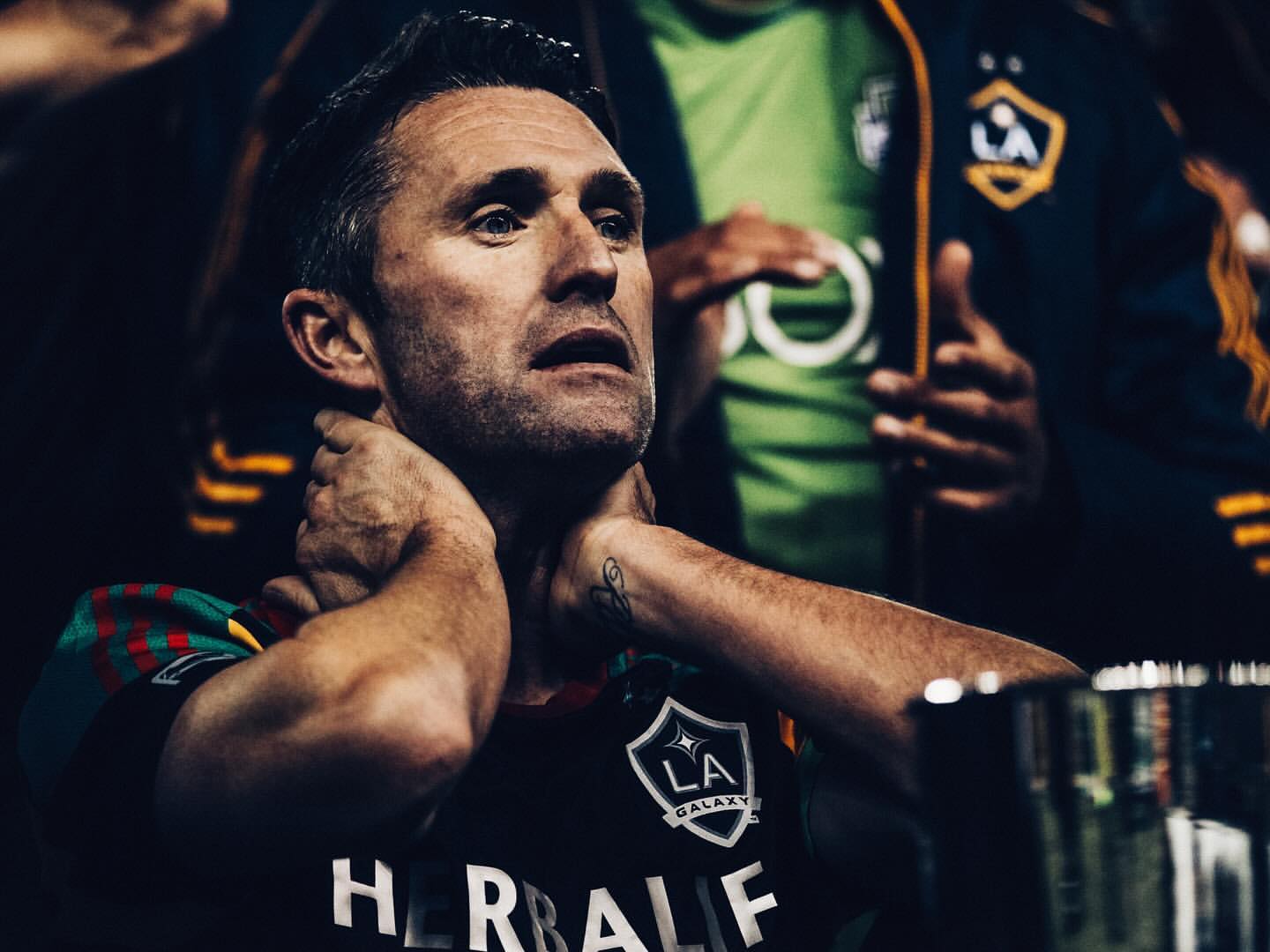Close-up of a middle-aged adult male in a sports jersey with his hands on his neck, looking up, with two other people in the background.