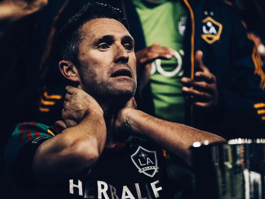 Close-up of a middle-aged adult male in a sports jersey with his hands on his neck, looking up, with two other people in the background.
