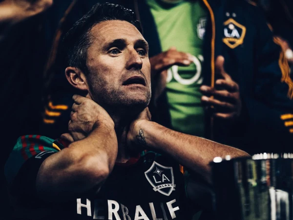 Close-up of a middle-aged adult male in a sports jersey with his hands on his neck, looking up, with two other people in the background.