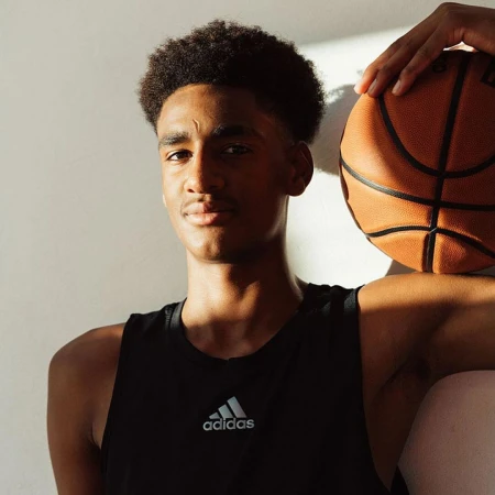 Portrait of a dark-skinned young adult male in a black tank top holding a basketball, leaning against a white wall in soft light.