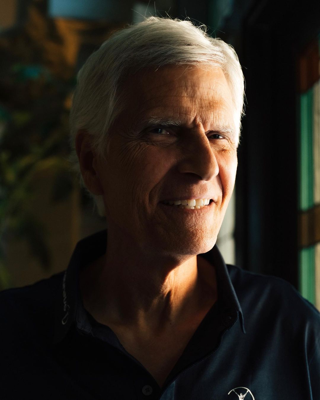Close-up portrait of a mature adult man with short gray hair, a slight smile, wearing a dark blue collared shirt indoors.