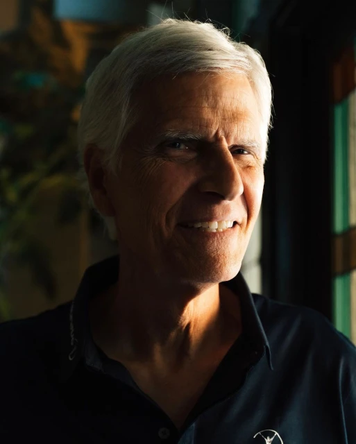Close-up portrait of a mature adult man with short gray hair, a slight smile, wearing a dark blue collared shirt indoors.