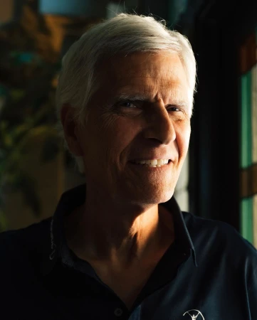 Close-up portrait of a mature adult man with short gray hair, a slight smile, wearing a dark blue collared shirt indoors.