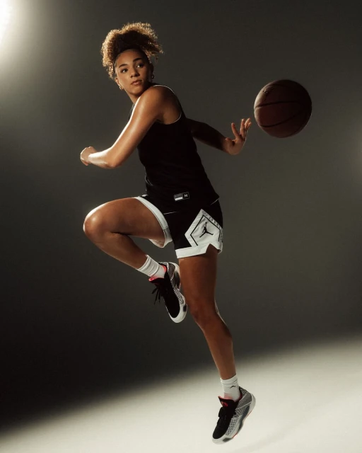 Athletic female basketball player in mid-air with a basketball, wearing a black jersey and shorts with sport shoes in studio lighting.