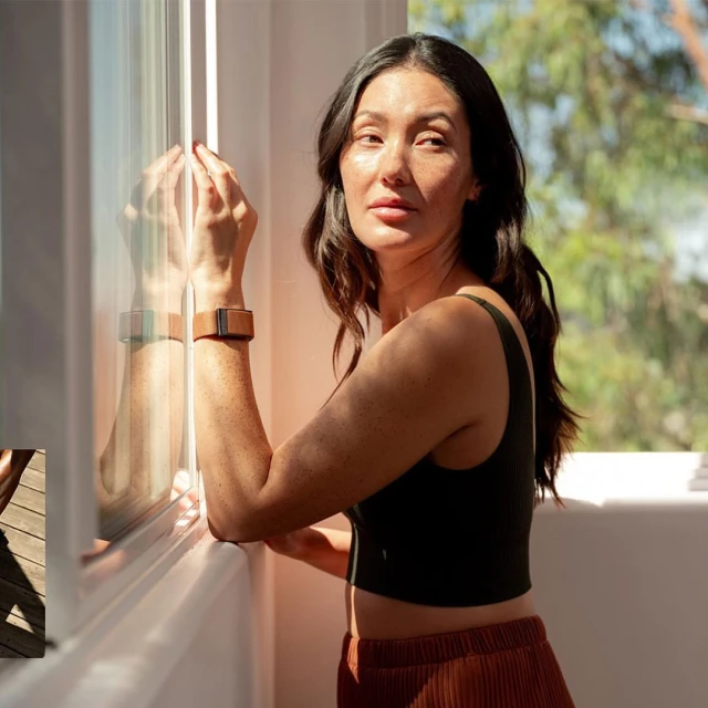 Portrait of a woman in a black tank top and rust-colored skirt standing near a window with sunlight casting shadows indoors.
