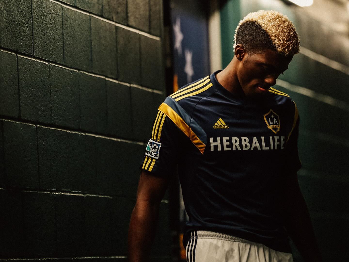 Male soccer player in team jersey posing in indoor setting with brick wall and dark lighting. Herbalife logo visible on jersey.