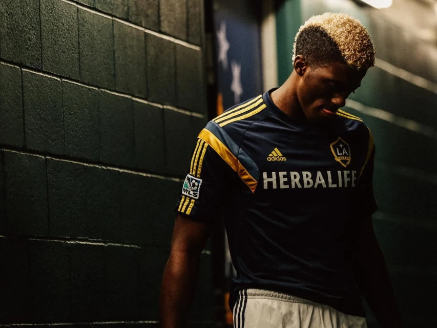 Male soccer player in team jersey posing in indoor setting with brick wall and dark lighting. Herbalife logo visible on jersey.