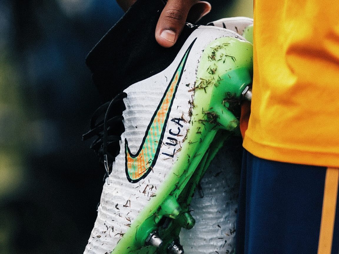 Close-up of soccer cleat in hand with the Nike logo and the name Luca on it; the shoe is worn and covered in grass.