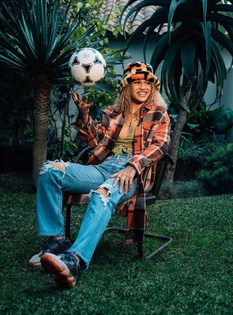 Young woman with curly hair sits in a chair outdoors, juggling a soccer ball in the air, wearing a colorful hat and jacket.