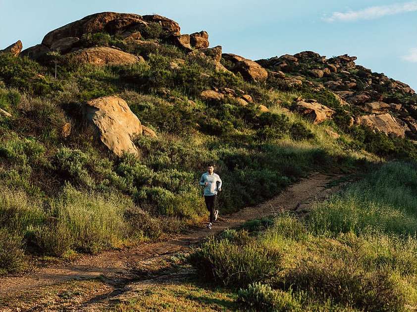 Adult male runs on a dirt path through a grassy, rocky landscape under a clear sky, capturing a moment of fitness and outdoor activity.
