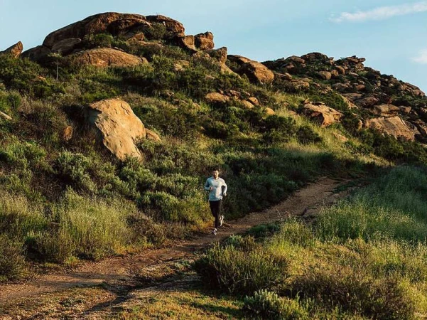 Adult male runs on a dirt path through a grassy, rocky landscape under a clear sky, capturing a moment of fitness and outdoor activity.