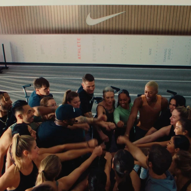 Group of diverse athletes in a huddle with hands together in a gym setting under the Nike logo. Teamwork and unity in sports.