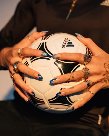 Close-up of hands holding a soccer ball, featuring rings, intricate manicured nails, and tattoos by photographer Hana Asano.