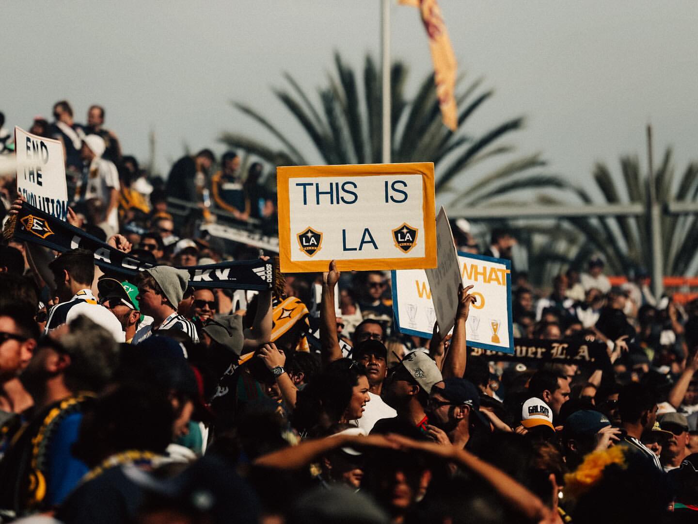 Crowd of soccer fans holding signs and wearing team colors and scarves supporting the LA Galaxy soccer team at an outdoor stadium.
