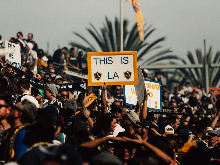 Crowd of soccer fans holding signs and wearing team colors and scarves supporting the LA Galaxy soccer team at an outdoor stadium.
