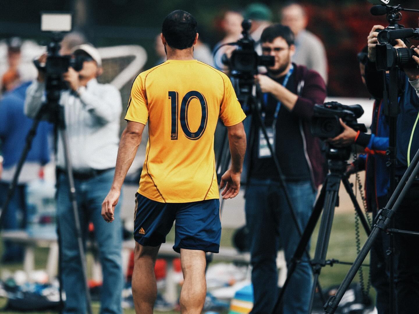 A soccer player in a yellow jersey with the number 10 walks away from the camera towards a group of news media and their filming equipment.