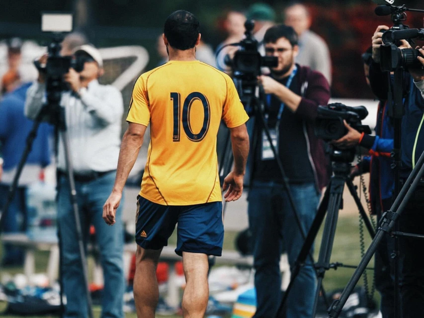 A soccer player in a yellow jersey with the number 10 walks away from the camera towards a group of news media and their filming equipment.