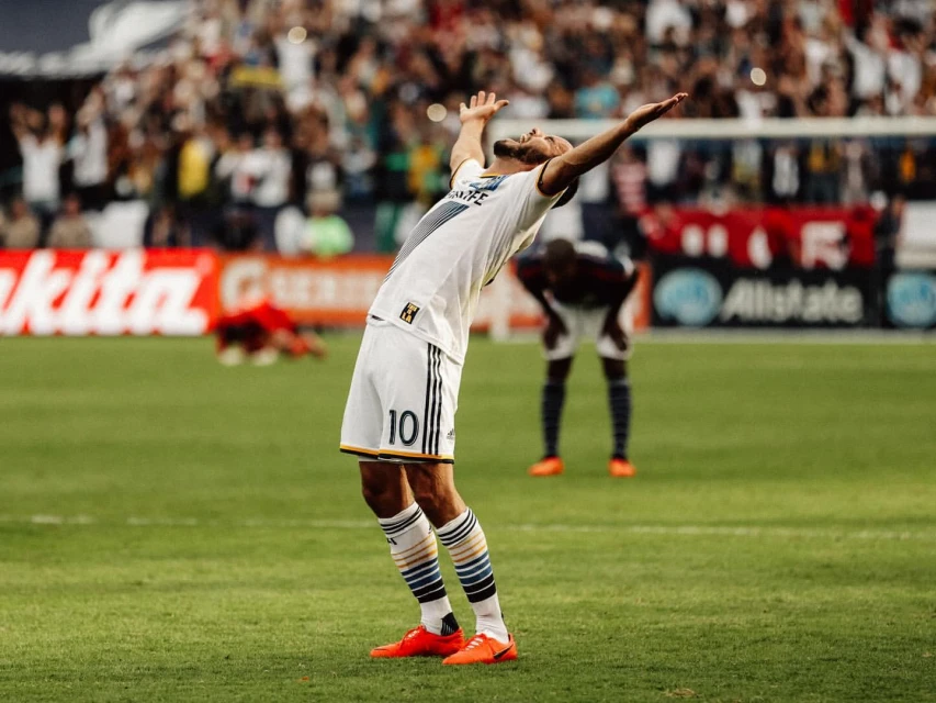 Soccer player celebrating a win on the field with arms raised in victory near other players and a crowd in the background.