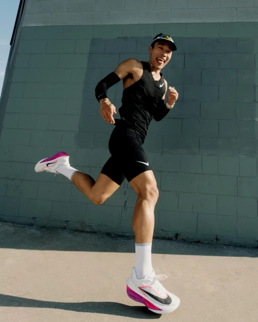 Asian man running in a black Nike tank top and matching shorts, with white and pink running shoes, outside in bright sunlight.