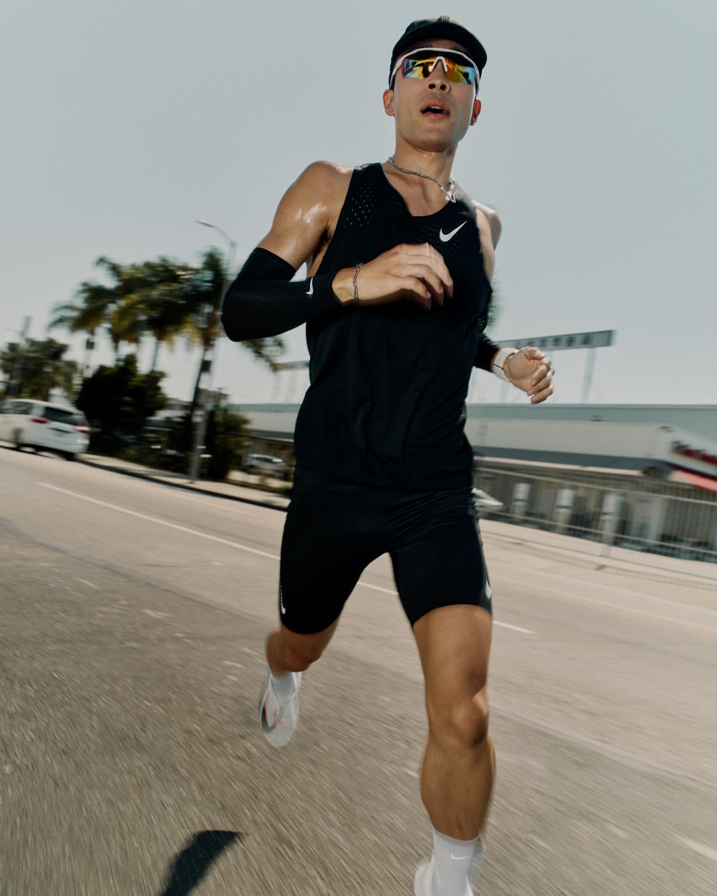A male runner in athletic wear and sunglasses is captured in motion on a city street, with buildings and palm trees in the background.